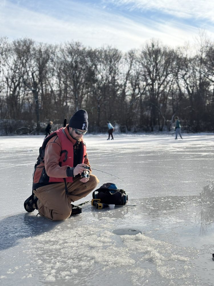 An angler fishes through the ice on long island. A game of pond hockey is being played in the background.